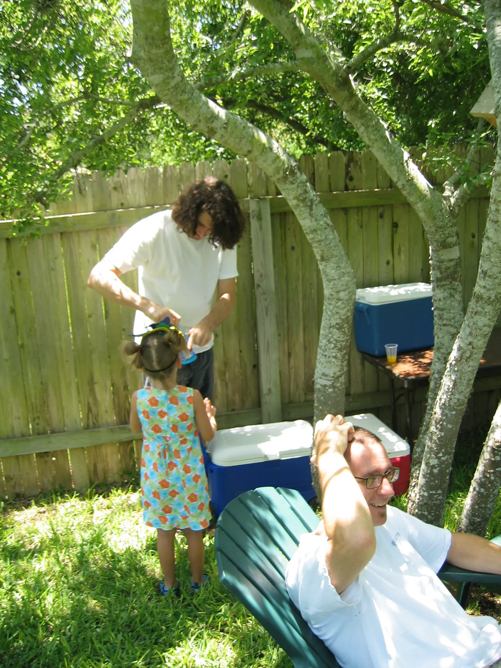 A little girl in a flowered dress getting her hair bow fixed while a man laughs in a lawn chair