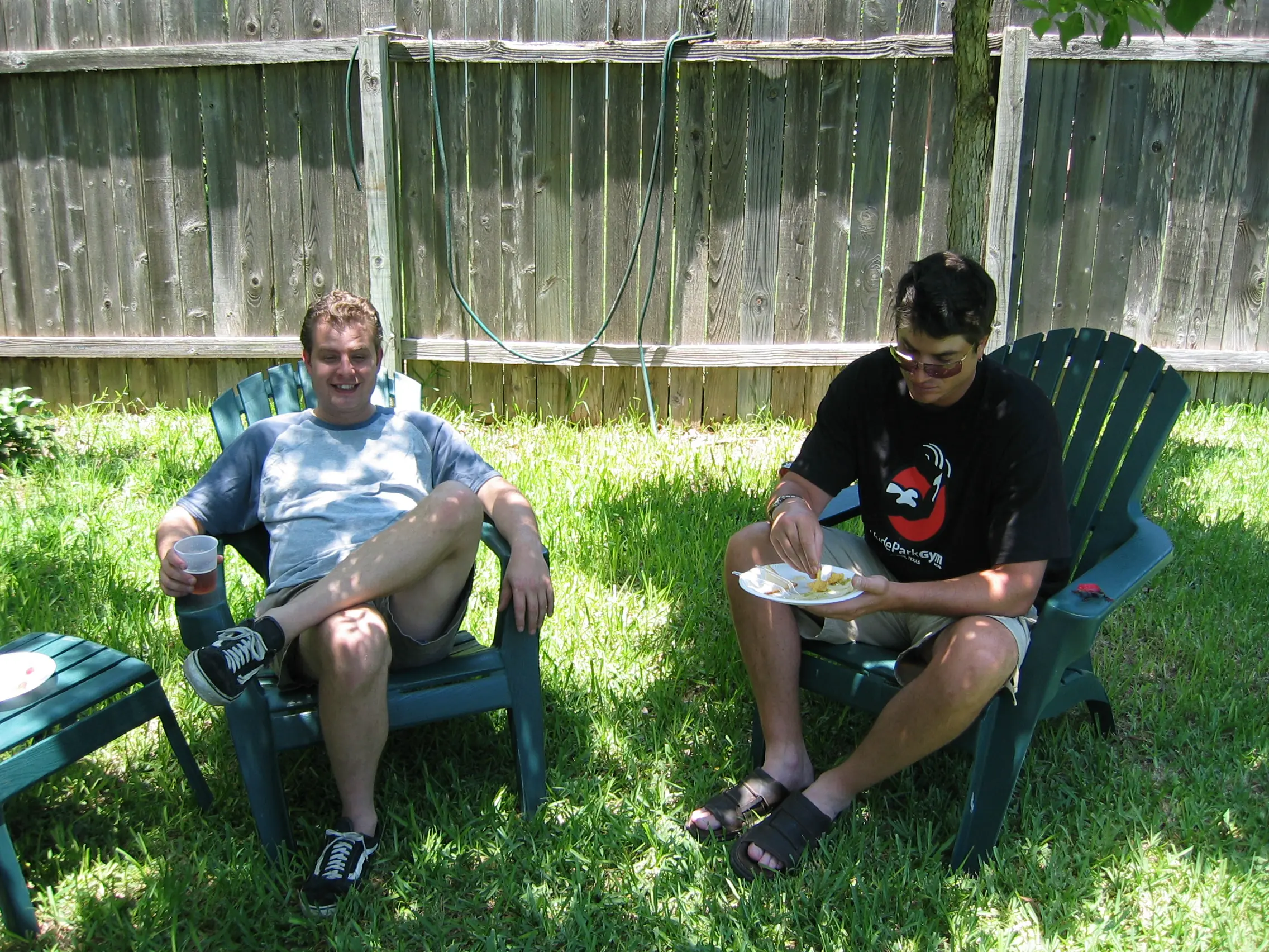 Two guys sitting in lawn chairs in the grass eating and drinking