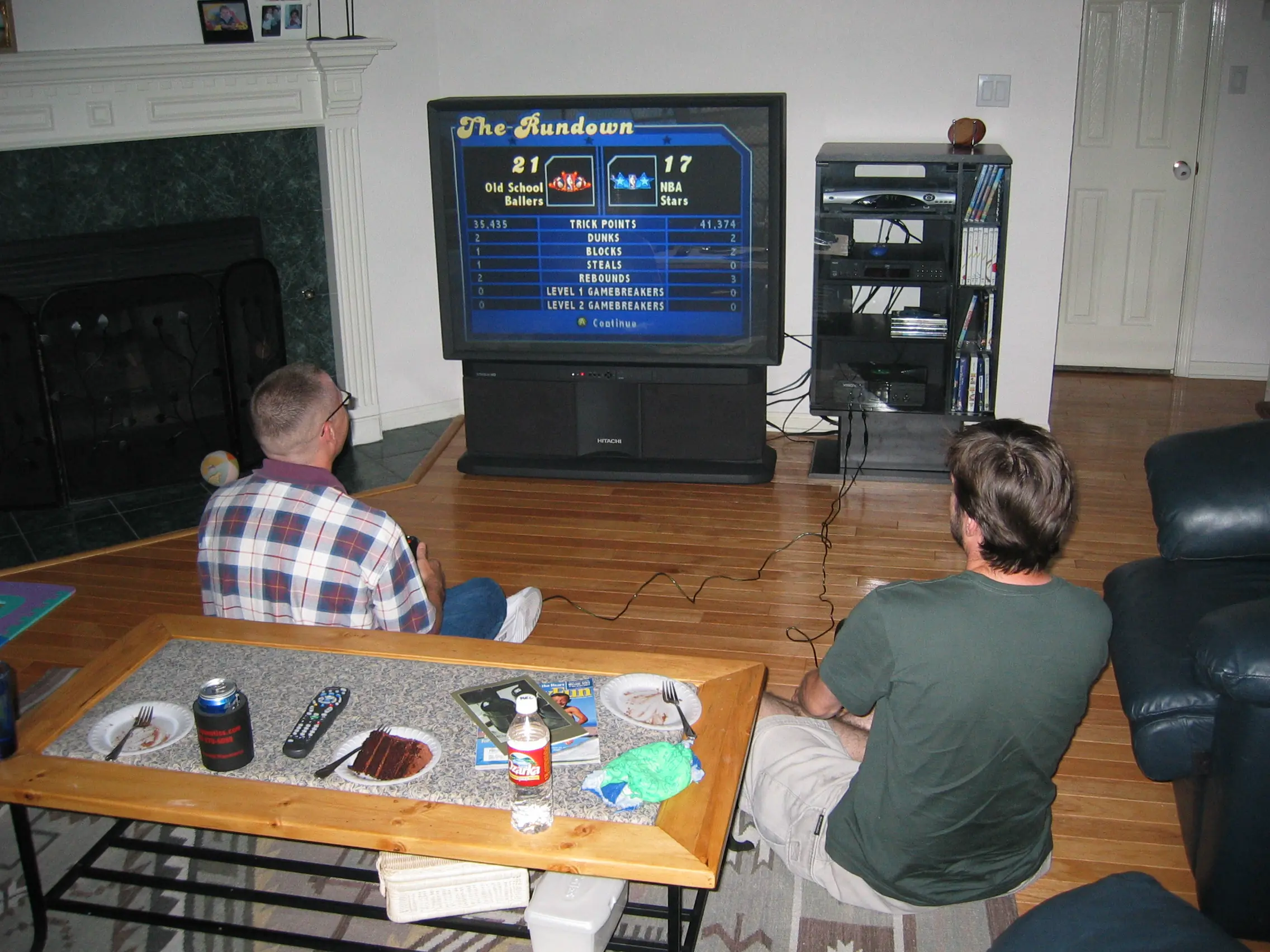 Two guys sitting on a living room floor playing NBA Street on a big rear-projection TV, with cake plates on the coffee table