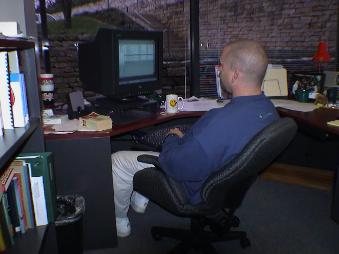 A developer in a blue pullover sitting at a corner desk working on a CRT monitor, with a smiley face mug and bookshelves nearby