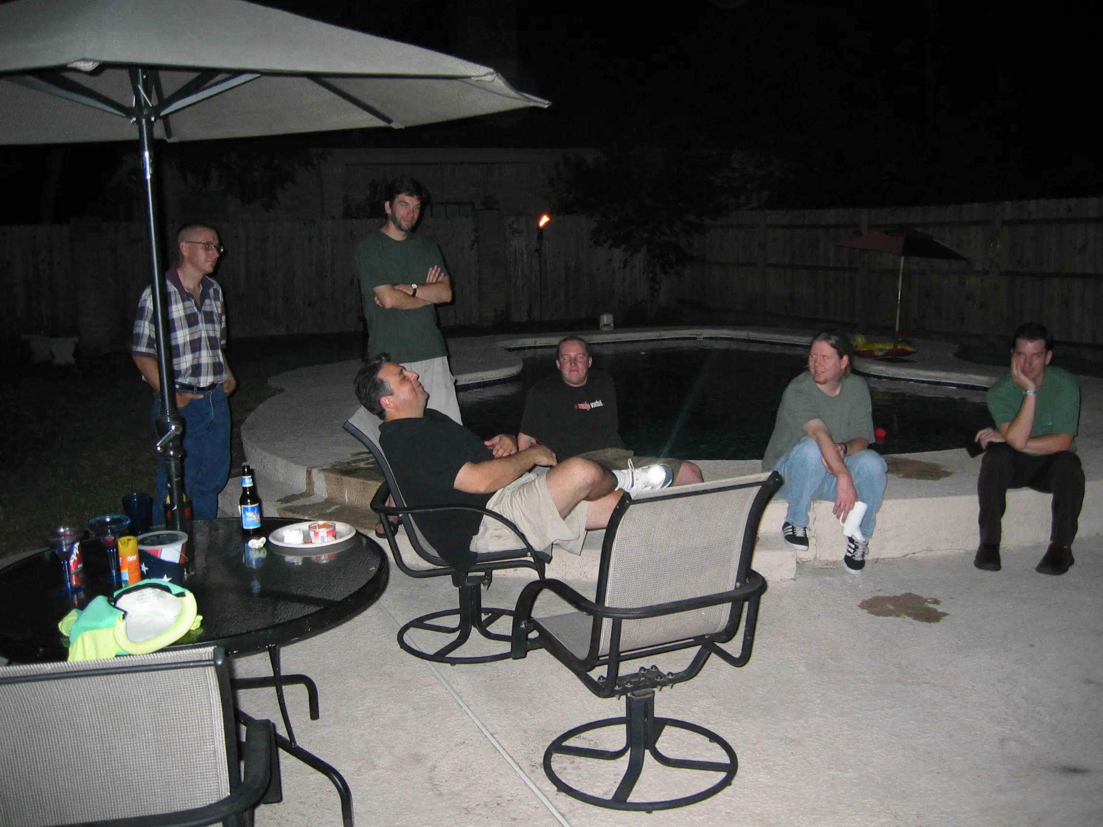 A group of friends hanging out around a backyard pool at night under a patio umbrella