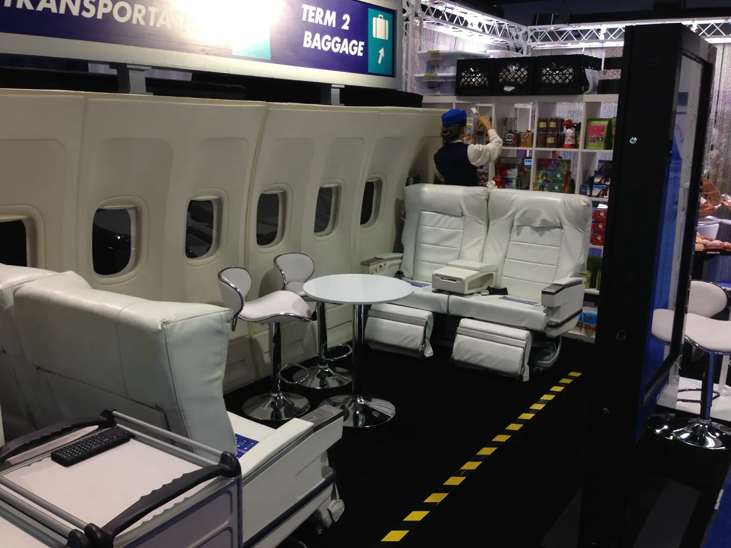 The movie-set airplane fuselage booth built for the tradeshow — white leather cabin seats, real aircraft windows, baggage-terminal signage, and a uniformed flight attendant stocking the duty-free retail shelf.